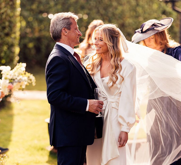 Man in blue suit holding champagne flute stands with bride in wedding dress with train and satin wrap top with veil in garden