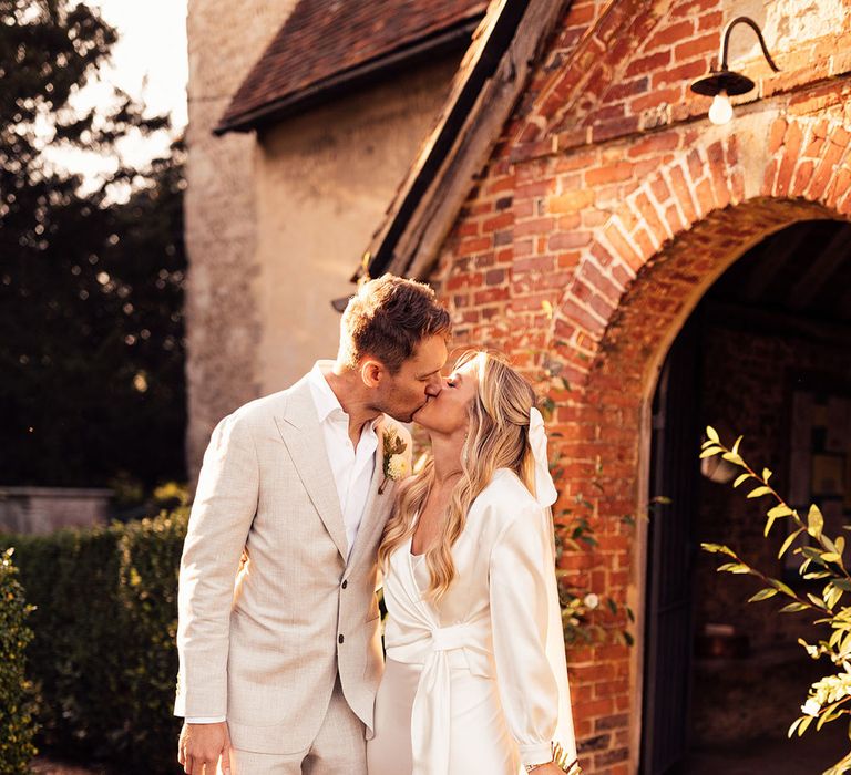 Bride in satin wedding dress and wrap top and groom in light grey suit kiss outside church after wedding ceremony