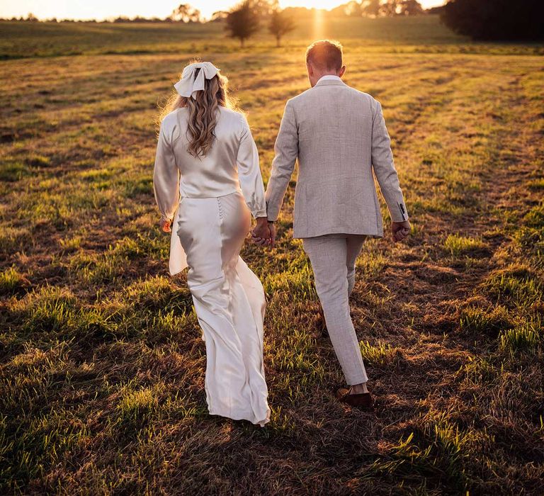 Bride with satin dress with train and hair bow walks through field during golden hour handing hands with groom in grey suit