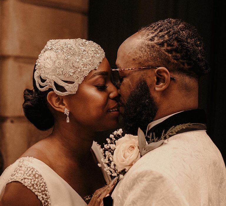 Black bride in an embellished cap headdress kissing her groom in a white tuxedo jacket 