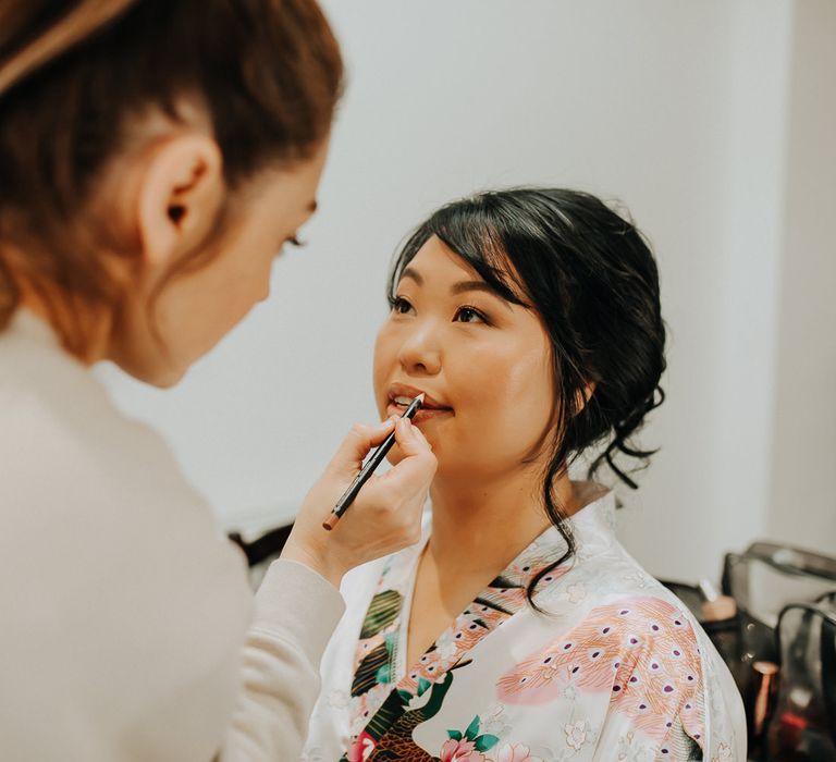 Bride in patterned silk kimono dressing gown sits having her lips lined before wedding 