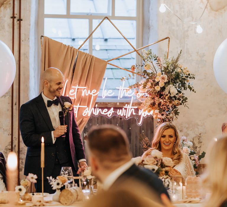 Groom in navy suit holds microphone whilst making speech during wedding reception with copper frame wedding arch and neon sign