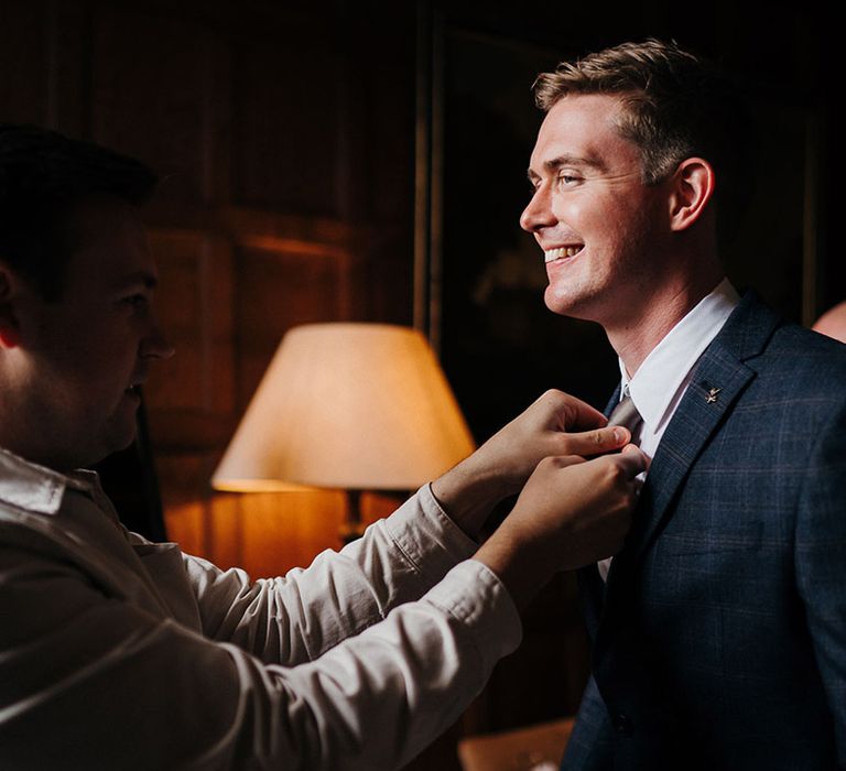 Groom gets his silver grey tie straightened in blue checkered suit for country house wedding