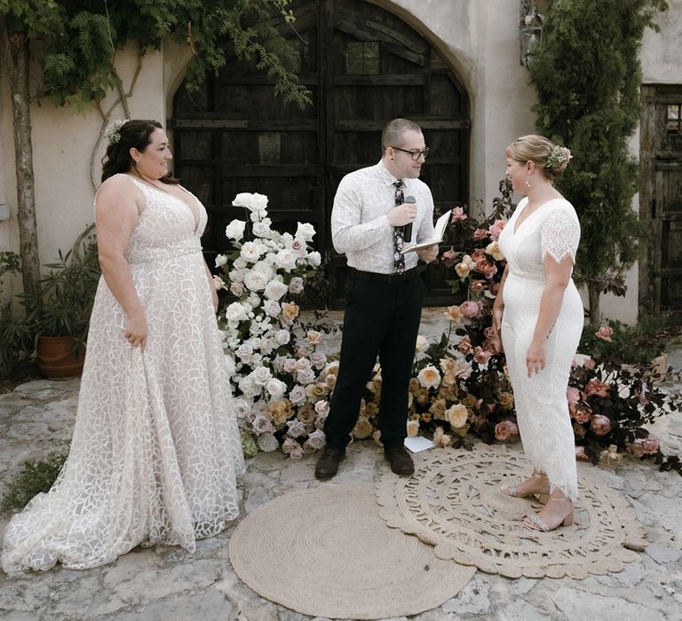 Bride wearing v-neck illusion wedding dress with geometric patterns with bride in lace jumpsuit with matching diamanté belts at the altar