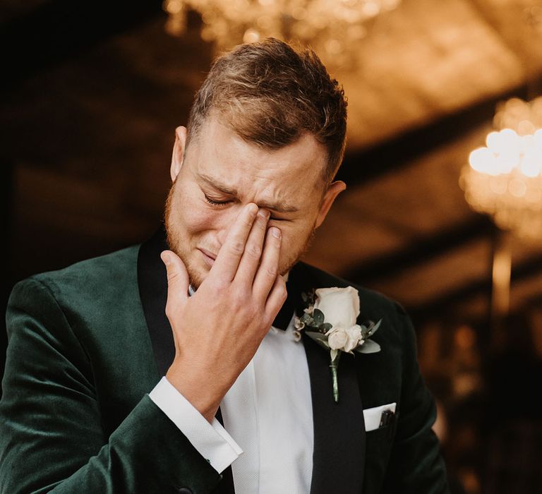 Groom in velvet green suit jacket and white rose boutonnière cries as he sees his bride 