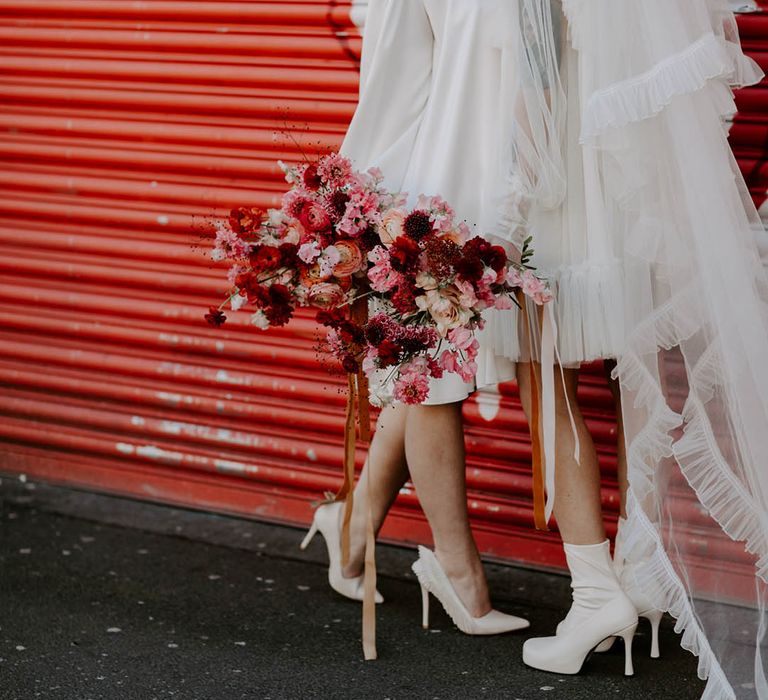 bride in short wedding dress long veil and boots wipering into her brides ear in a short dress and berret hat as she holds a red and pink bouquet 