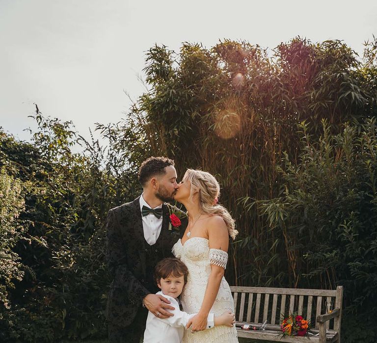 Groom in black tie with patterned tuxedo and dark green bow tie holding onto son in black tie and bride in boho lace strapless wedding dress 