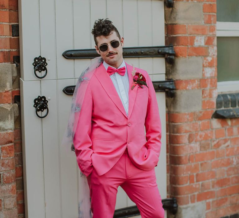 Stylish groom in a pink two-piece suit with bow tie, sunglasses and embroidered wedding veil 