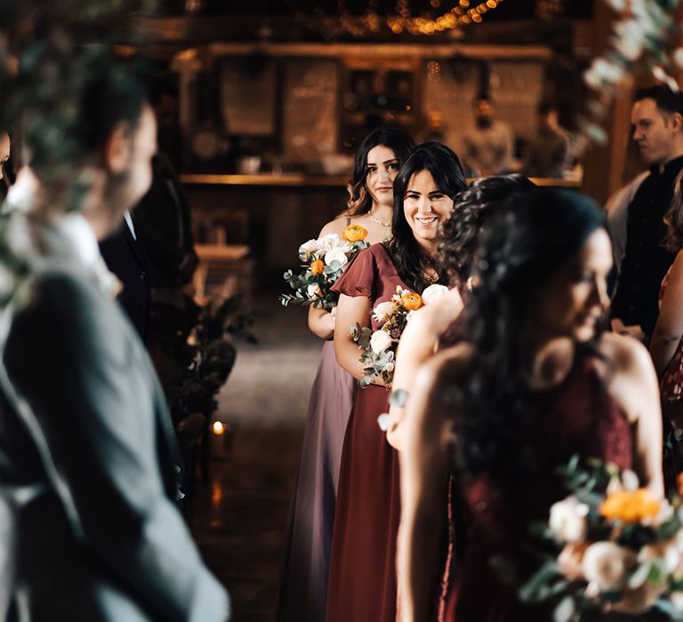 Bridesmaids in deep red, burgundy and pink dresses walk down the aisle