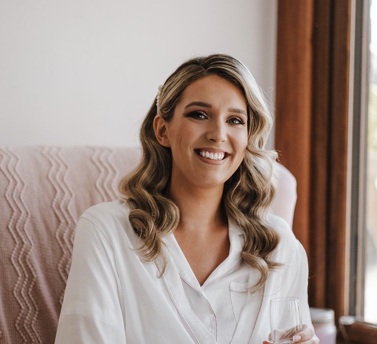 Bride wearing pearl hair clip and white robe holding champagne glass with French tip wedding nails 