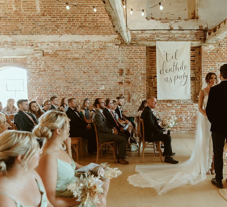 Bride and groom stand at the altar with wedding drapes and candles for altar decor with white banner reading 'til death do us party'