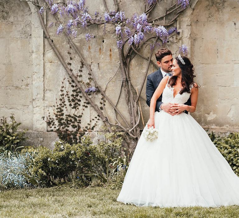 Groom hugs the bride from behind wearing a Pronovias princess style wedding dress with tiara crown in front of Kirtlington Park House