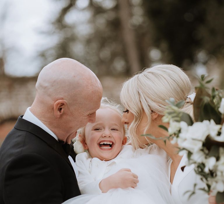 Bride and groom hold their daughter inbetween them as she giggles in tulle white dress and pearl cardigan 