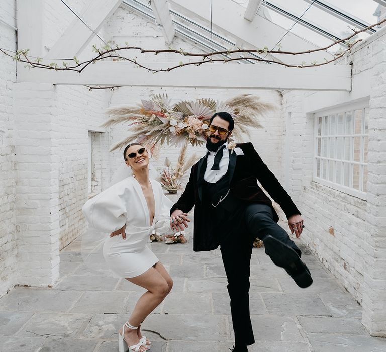 Bride in a short wedding dress with long sleeves, platform shoes and sunglasses holding hands with her groom in a tuxedo in the summer house at Garthmyl Hall 