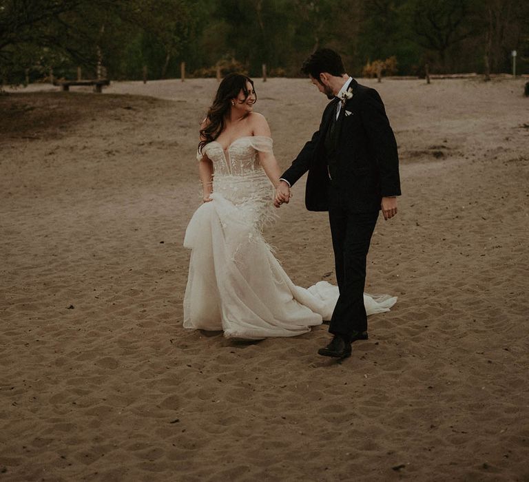 Bride and groom walk onto a sandy beach holding hands 