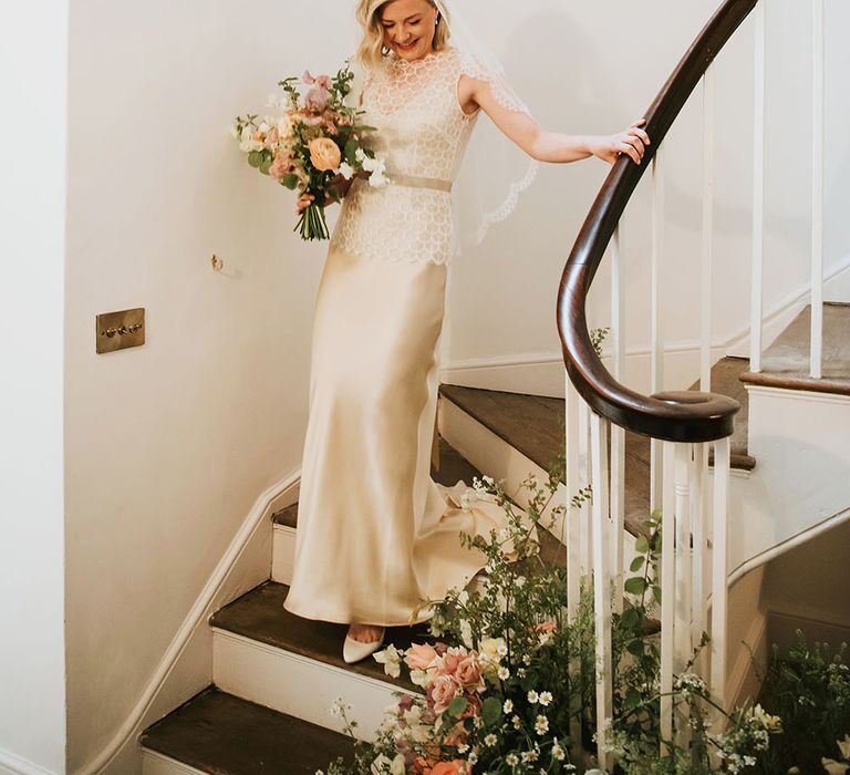 Bride walks down staircase at Aswarby Rectory on the morning of her wedding day in gold silk wedding gown complete with pastel floral bouquet