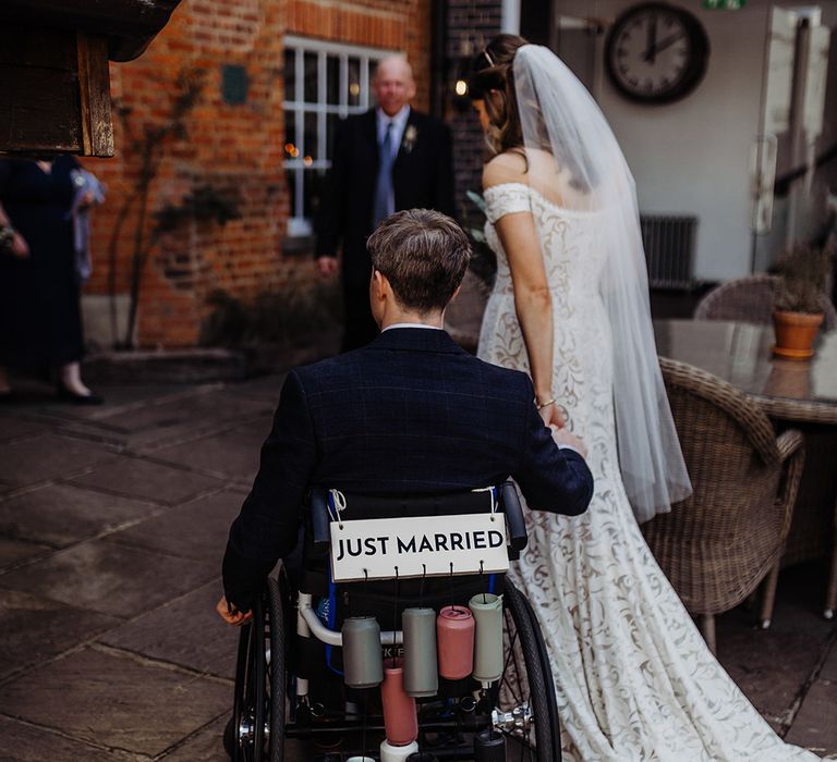 Groom in a blue suit in a wheelchair with 'Just Married' sign attached and colourful painted cans on string 