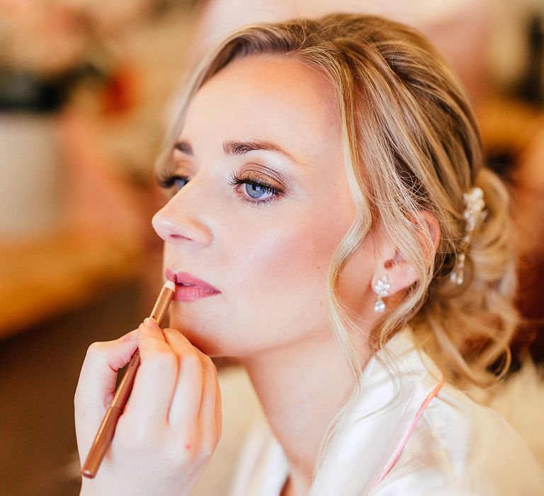 Bride gets her makeup done for her wedding day wearing white satin pyjamas with her hair in an updo with pretty hair pins 