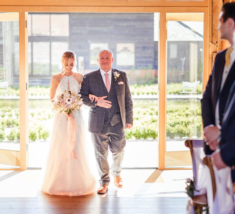 Father of the bride in grey suit with a pink tie walks the bride in a tulle wedding dress down the aisle 