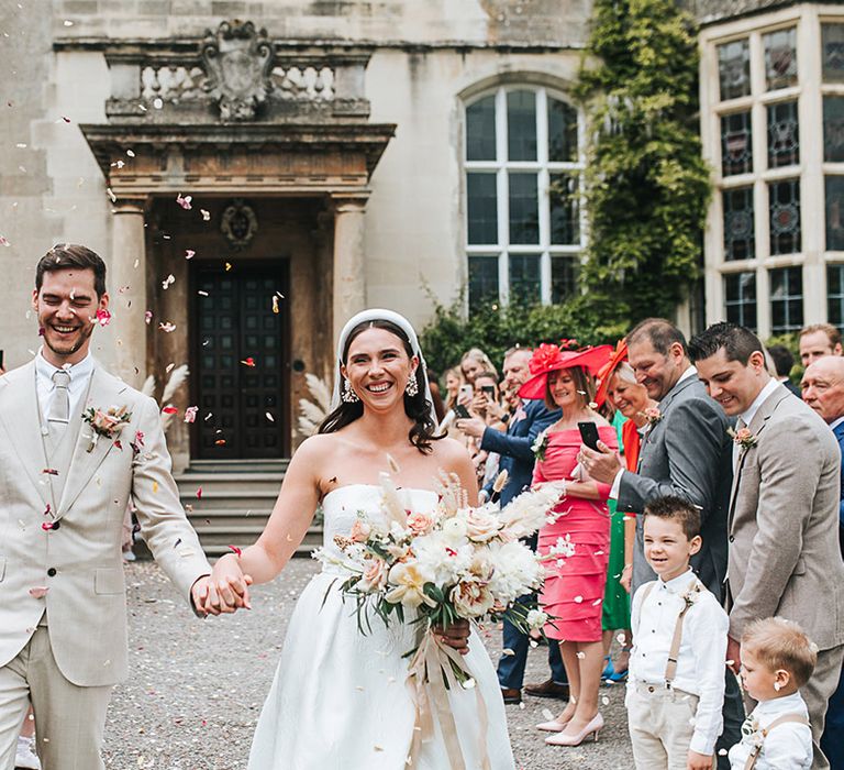 Groom in all neutral beige wedding suit smiles widely with the bride as they have a confetti moment 