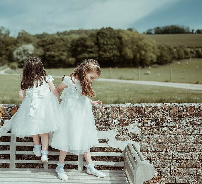 Flower girls in white dresses with a satin ribbon standing on a wooden bench