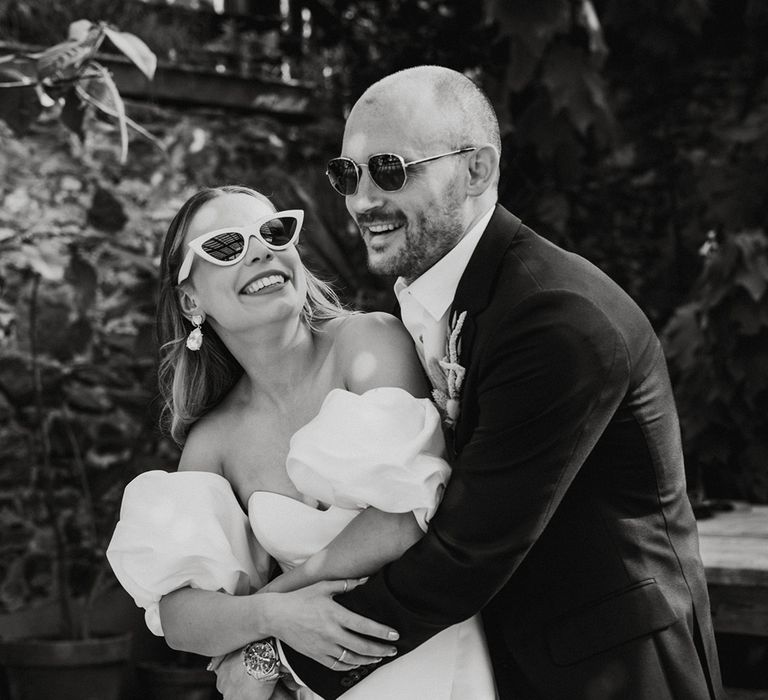 Groom wearing sunglasses hugs the bride from behind in a puff sleeve wedding dress with white sunglasses