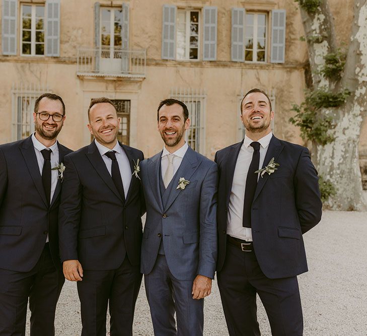 Groom wearing three-piece blue suit stands with his groomsmen in black suits and white shirts with floral buttonholes