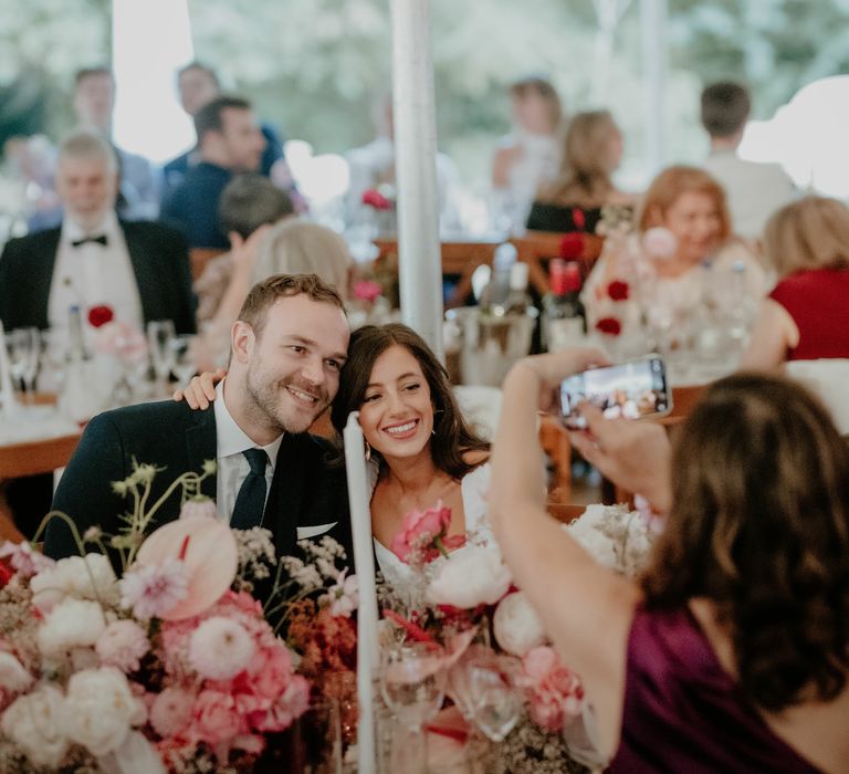 Bride & groom pose for photo whilst sat at wooden banquet tables surrounded by white and pink flower arrangements 