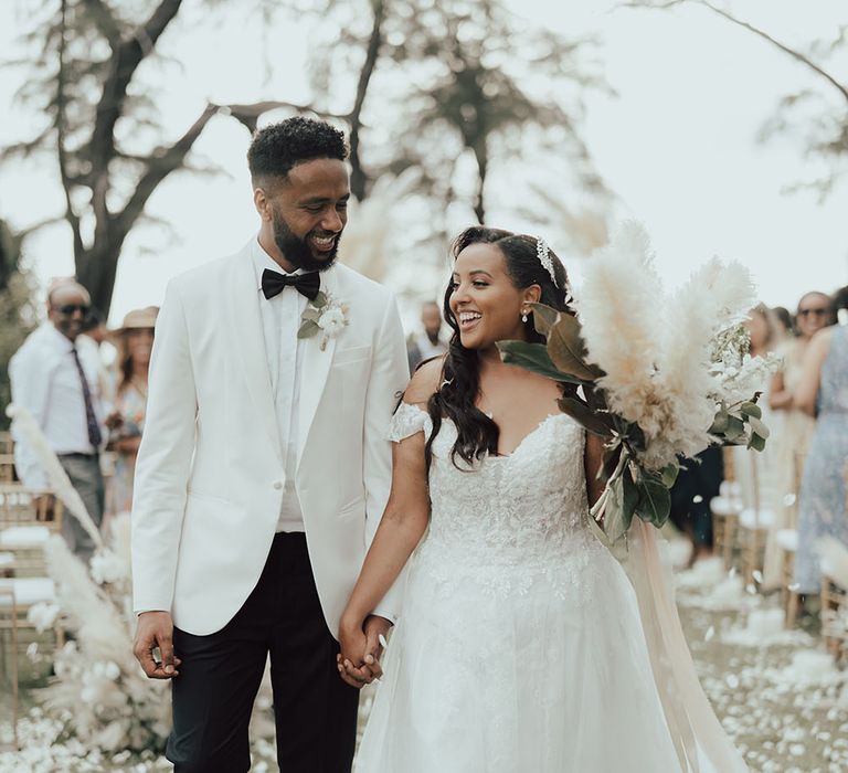 Groom wears black-tie and white suit jacket whilst walking with his bride in off-the-shoulder fitted lace wedding dress 