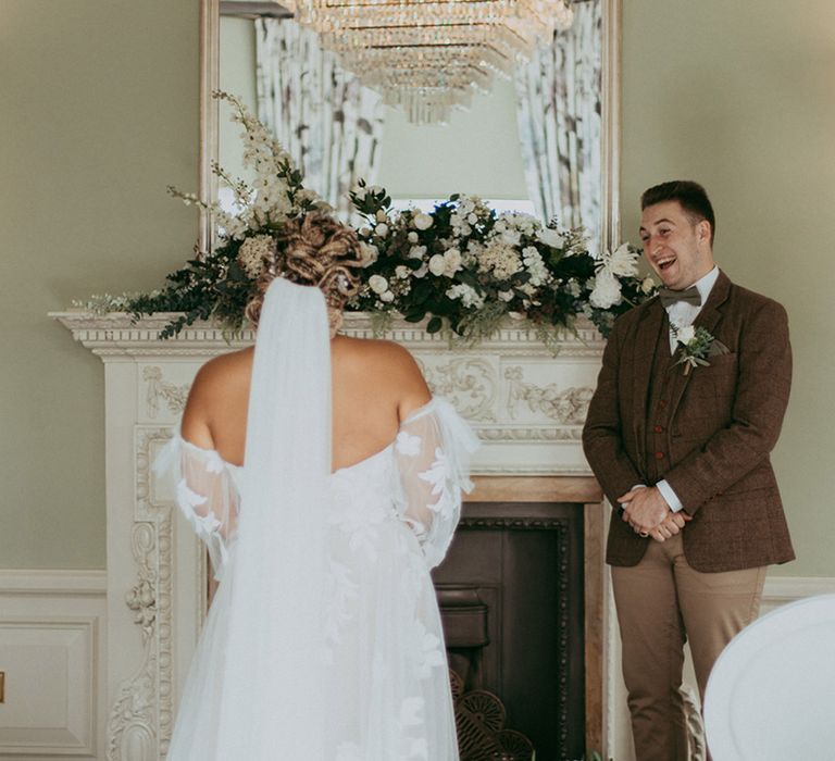 Groom in a brown checkered suit smiles widely as the bride in a sweetheart sheer puff sleeve wedding dress walks down the aisle 