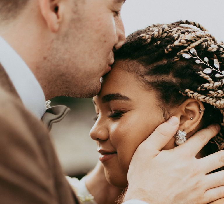 Groom kisses the bride on the forehead who wears pearl earrings and has her hair in blond braids with a silver hair accessory 