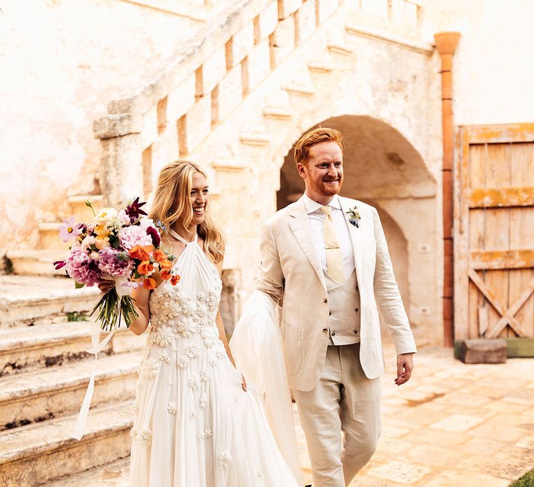 Groom in a three piece cream wedding suit walking with the bride in a halter neck flower wedding dress 