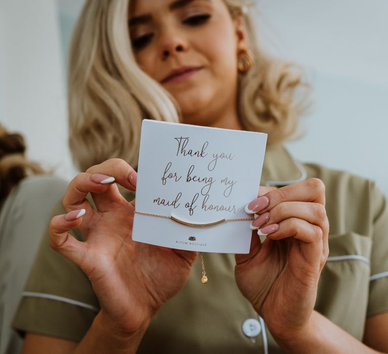 Maid of honour in sage green pyjama shirt opening wedding favour gold bracelet that says "Thank you for being my maid of honour"