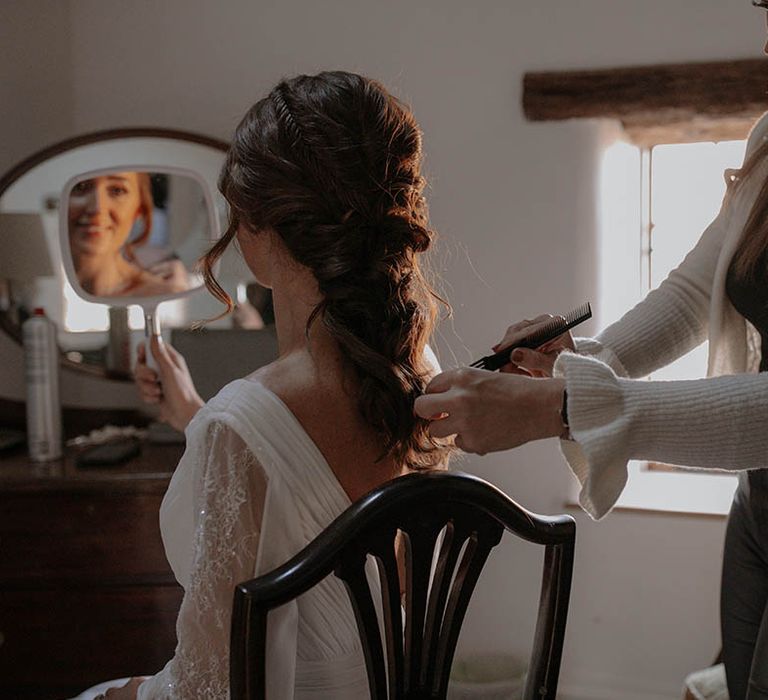 Bride gets her hair styled into a loose messy braided hairstyle in a long sleeve wedding dress 