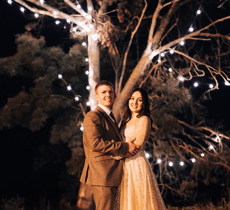 Bride & groom embrace outdoors in front of tree covered in festoon lighting 