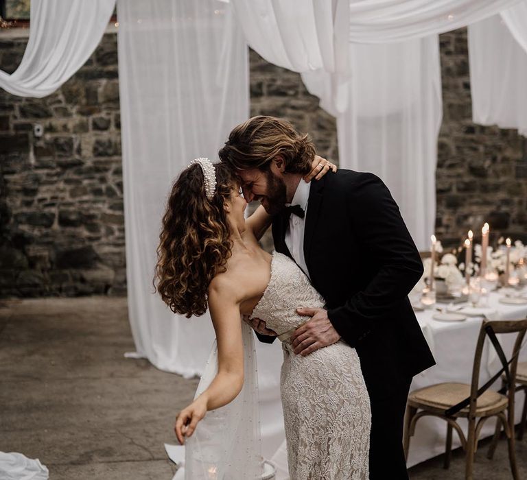 Groom in black tux and bride in sleeveless lace wedding dress with pearl details, pearl headband and silver belt dancing kissing and dancing