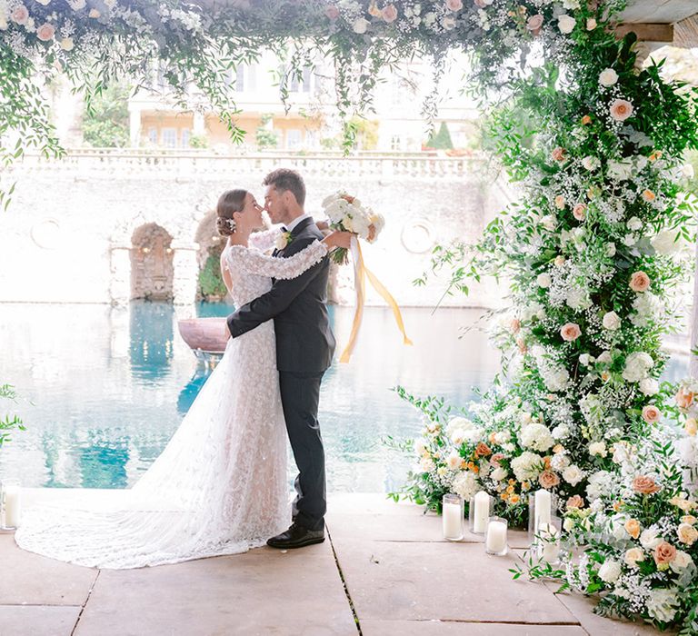 Bride and groom lean in to share a kiss surrounded by cream and yellow wedding flowers for their outdoor ceremony 