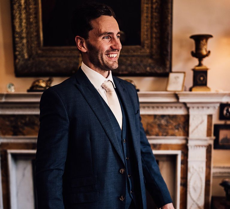 The groom in three piece navy blue suit and pale pink tie smiling as he gets ready for his wedding day 