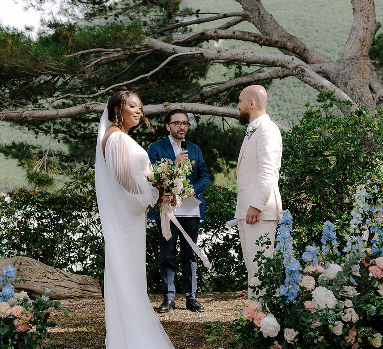 Bride smiles whilst the wedding celebrant speaks, surrounded by trees and beautiful floral installations