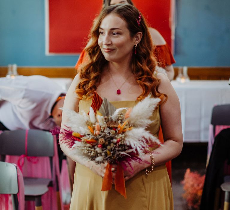 Bridesmaid in mustard sleeveless bridesmaid dress holding autumnal coloured bouquet with white pampas grass and an orange ribbon walking down the aisle