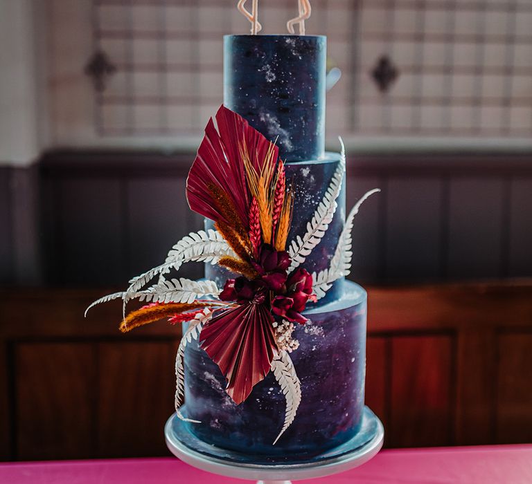 Deep blue three tiered wedding cake with white paint splatters and autumnal red flower decorations on the side and flamingos wedding cake topper