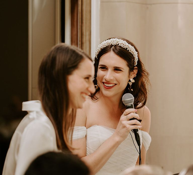 The bride wearing a pearl headband and statement earrings stands with a microphone to deliver a speech 