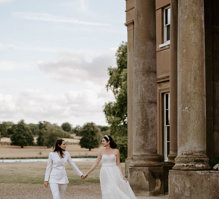 The bride in an off the shoulder wedding dress and bride in suit walk holding hands at the Wilderness Reserve wedding venue 