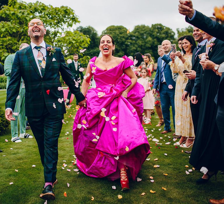 Groom in dark forest green tartan suit with patterned tie, white pocket square and pink and green boutonniere and bride in off the shoulder front ruching pink Vivienne Westwood wedding dress and dark red strappy heels doing flower petal confetti walk 