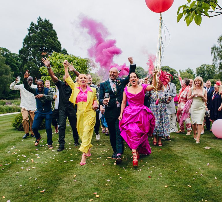 Groom in dark forest green tartan suit with patterned tie, white pocket square and pink and green boutonniere and bride in Vivienne Westwood off the shoulder front ruching pink wedding dress and Vivienne Westwood elevated red patent leather platform heels with shoe-string fastening leading colourful wedding party across Middleton Lodge with pink smoke bomb and balloons with gold streamers