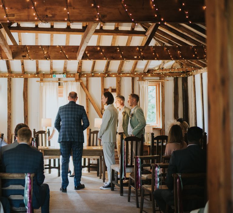 Groom in neutral coloured ASOS suit and high-top converse waiting at the end of alter at The Reid Rooms with fairy lights woven into the beams