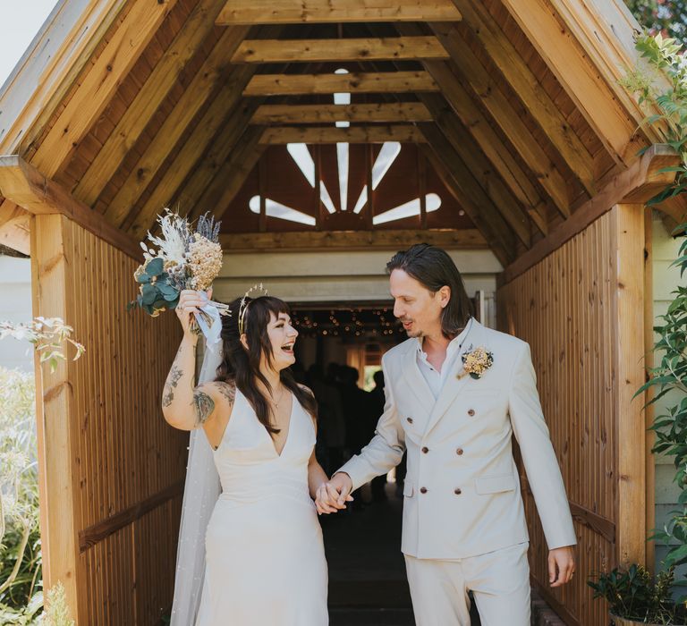 Groom in double-breasted ASOS suit and black high-top converse with dried flower and foliage boutonniere holding hands with bride in silk v neck ASOS wedding dress holding dried floral bouquet 