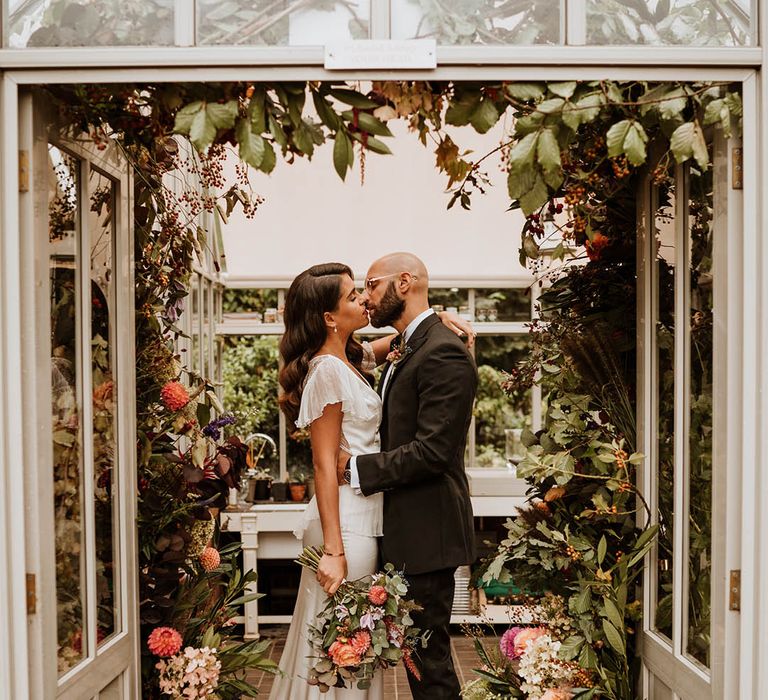 Bride in v-neck satin wedding dress with sheer butterfly sleeves and ruffled layer on top kissing groom in classic black tuxedo with hand-painted silk cravat and handkerchief at Le Manoir Aux Quat Saisons glasshouse venue