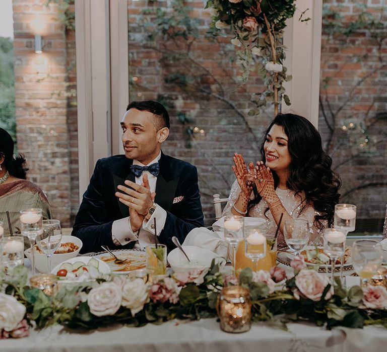 Pastel pink roses and green foliage line front of the table as bride & groom clap during wedding speeches