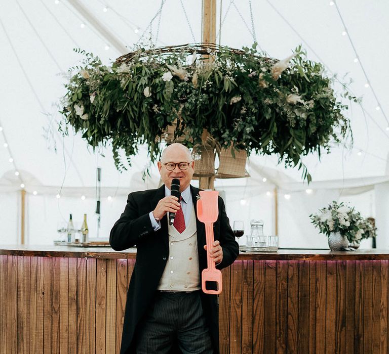 Wedding guest gives speech in marquee beside green foliage floral installation and wooden bar